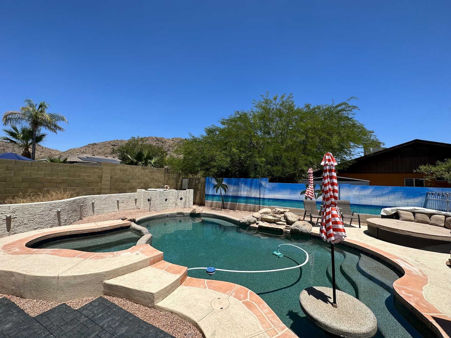 Heated pool and raised hot tub at South Mountain Oasis, with a red-and-white striped umbrella, painted beach-mural walls and South Mountain behind under a clear sky