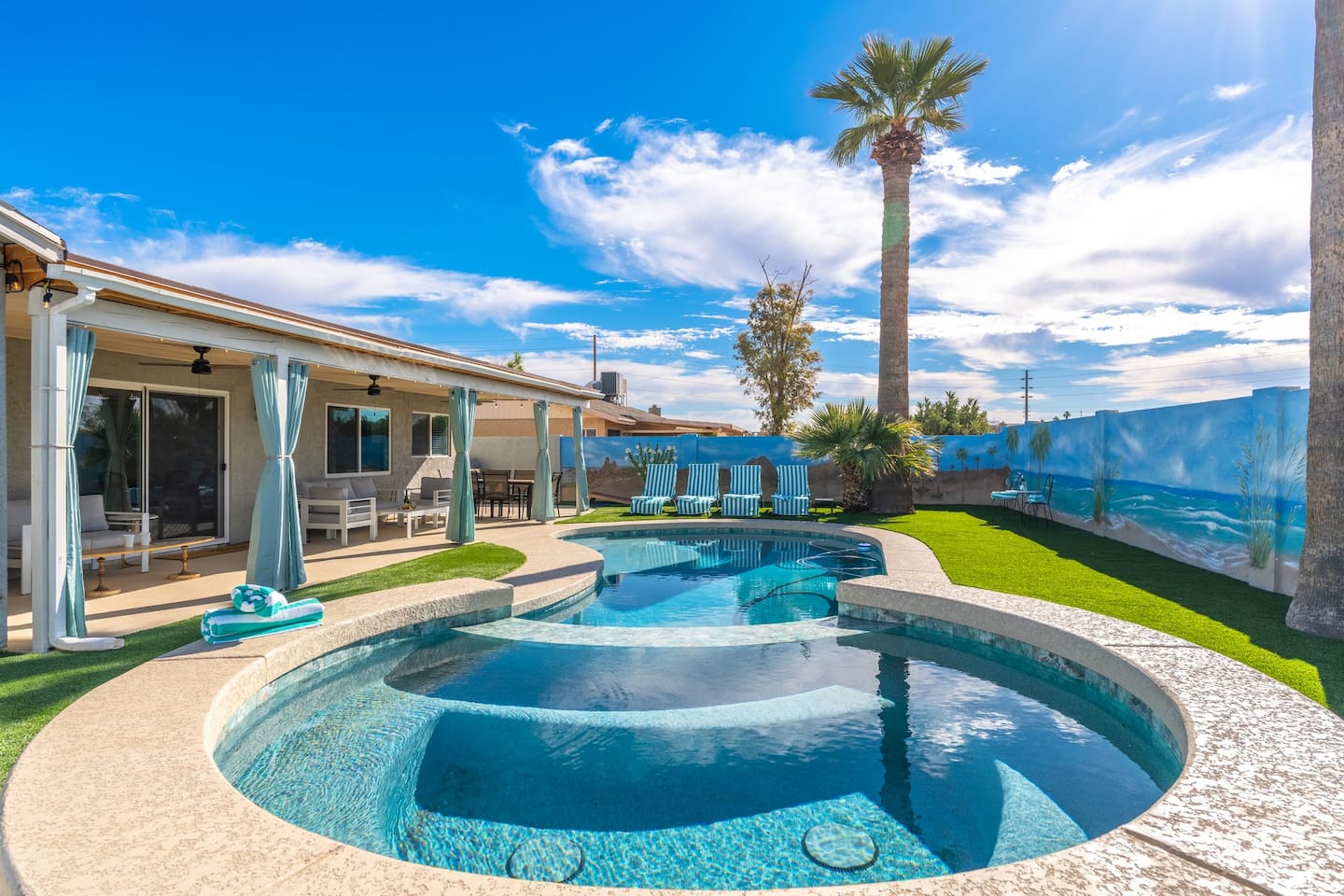 The round heated spa spilling into the pool at South Mountain Lagoon, with a covered patio, striped loungers on the turf, a tall palm and a painted beach-mural wall