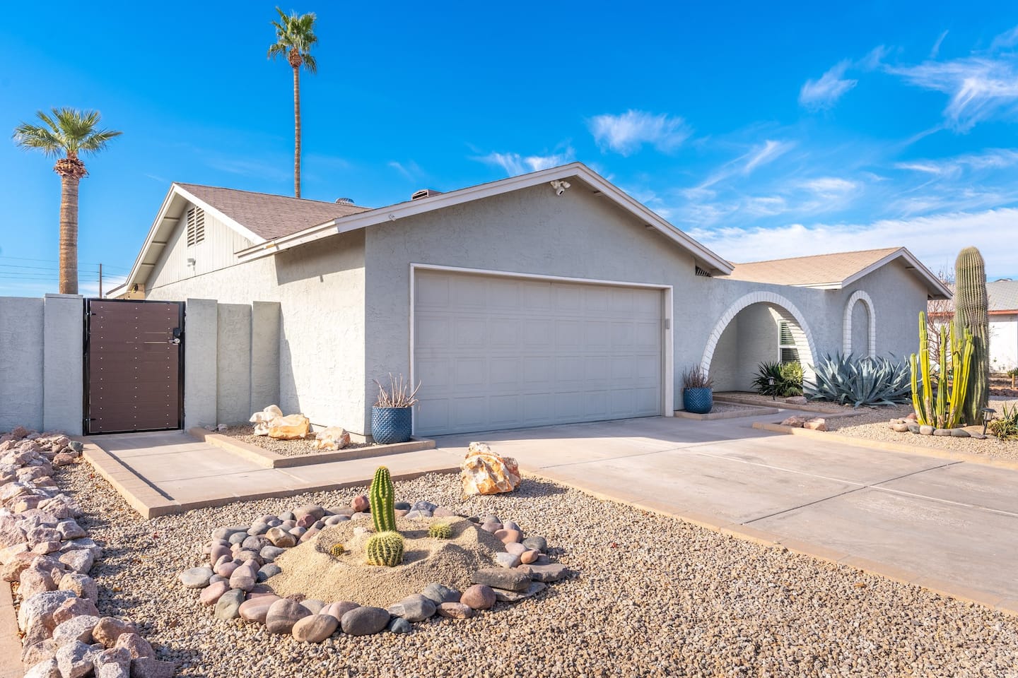 Street view of South Mountain Lagoon: a single-story stucco home with a two-car garage, palms behind and a bright sky