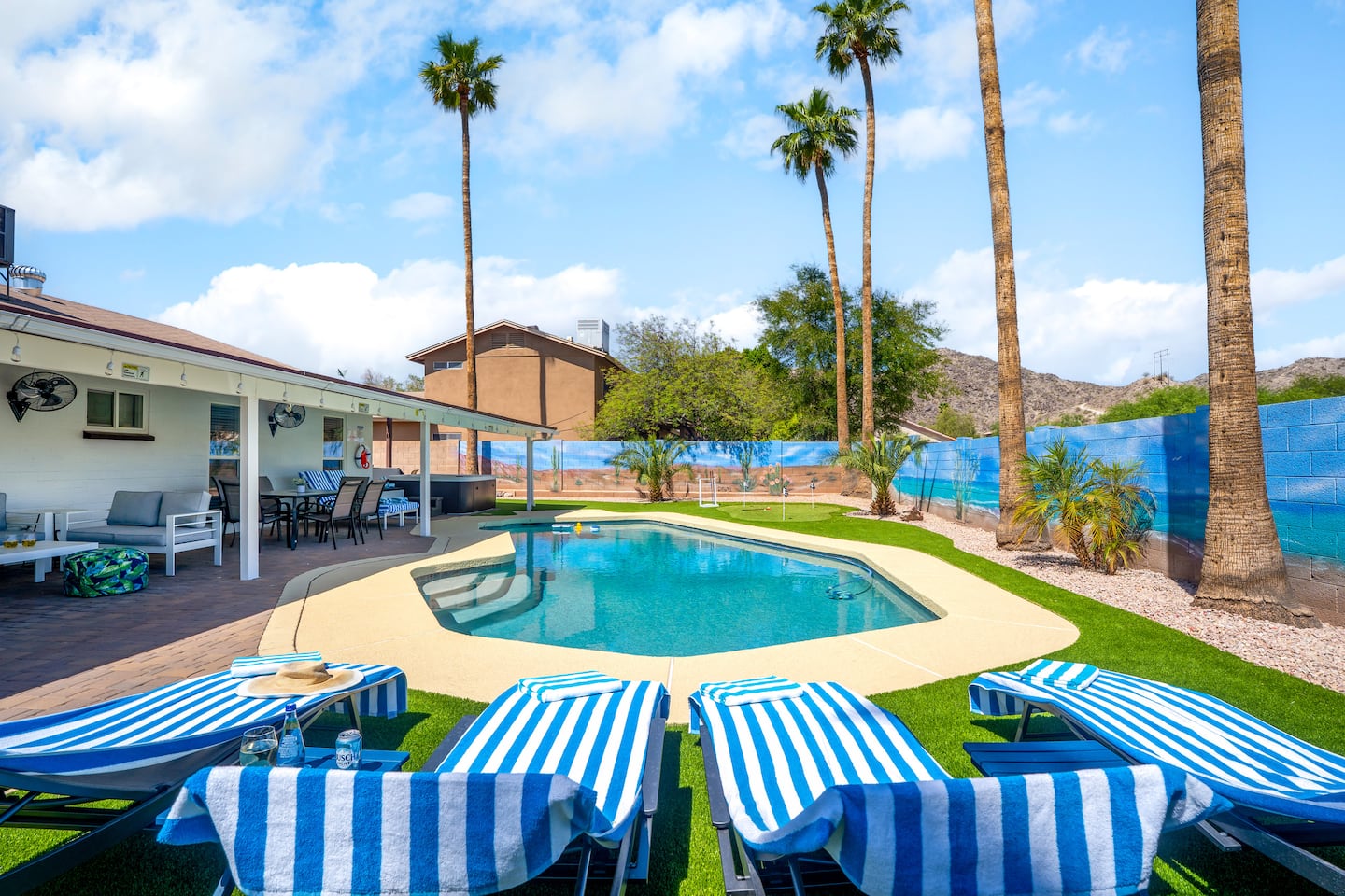 The backyard pool at South Mountain Vista under tall palms and a blue sky, ringed by blue-and-white striped loungers, with painted beach murals on the walls and the mountains behind
