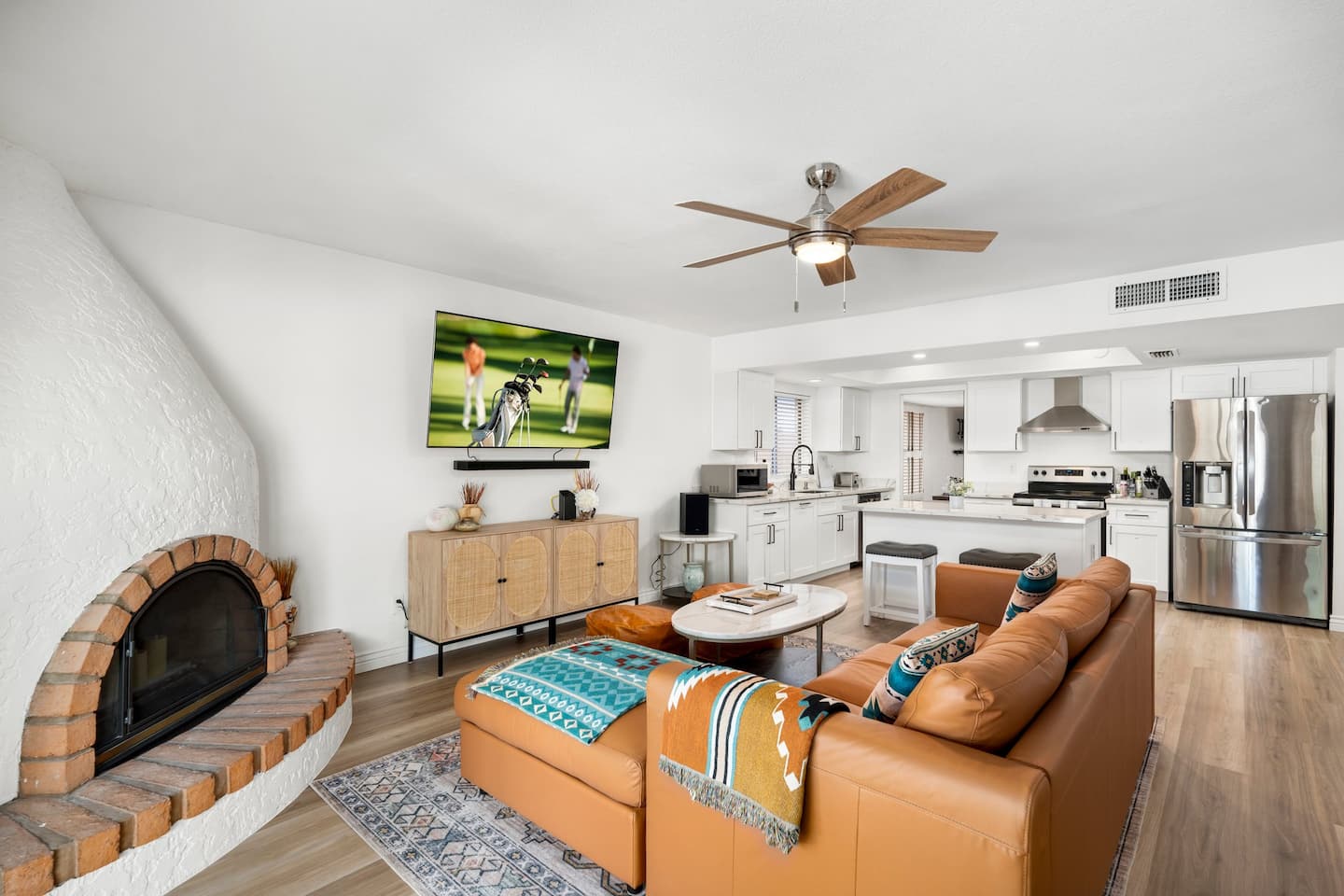 Living room at South Mountain Oasis: a cognac leather sectional, a white beehive fireplace with a brick hearth, and a wall-mounted TV open to the kitchen
